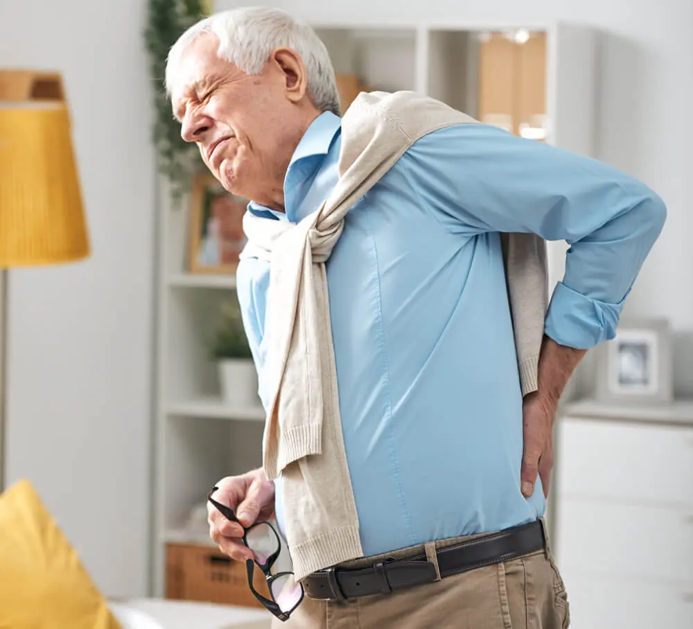 Älterer Mann zwischen 60 und 70 Jahren, der sich mit schmerzverzerrtem Gesicht den Rücken hält, als mögliches Symptom bei Prostatakrebs.